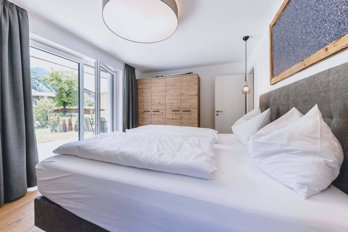 A bedroom with a large bed, a wooden wardrobe, a grey headboard, and a view of the outdoors.