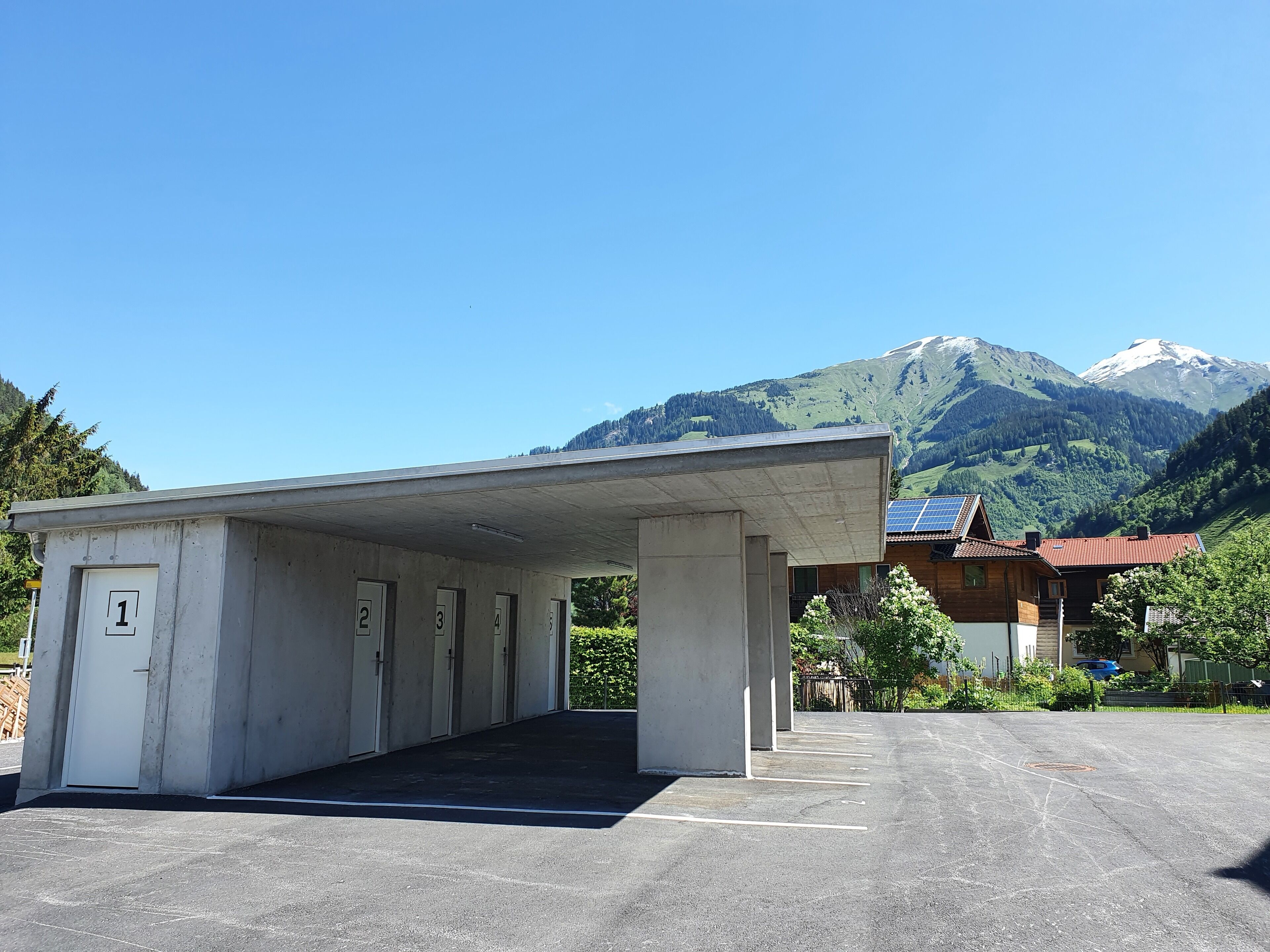 Ein Parkplatz mit nummerierten Türen, die zu einem Gebäude führen, vor einer Bergkulisse unter blauem Himmel.
