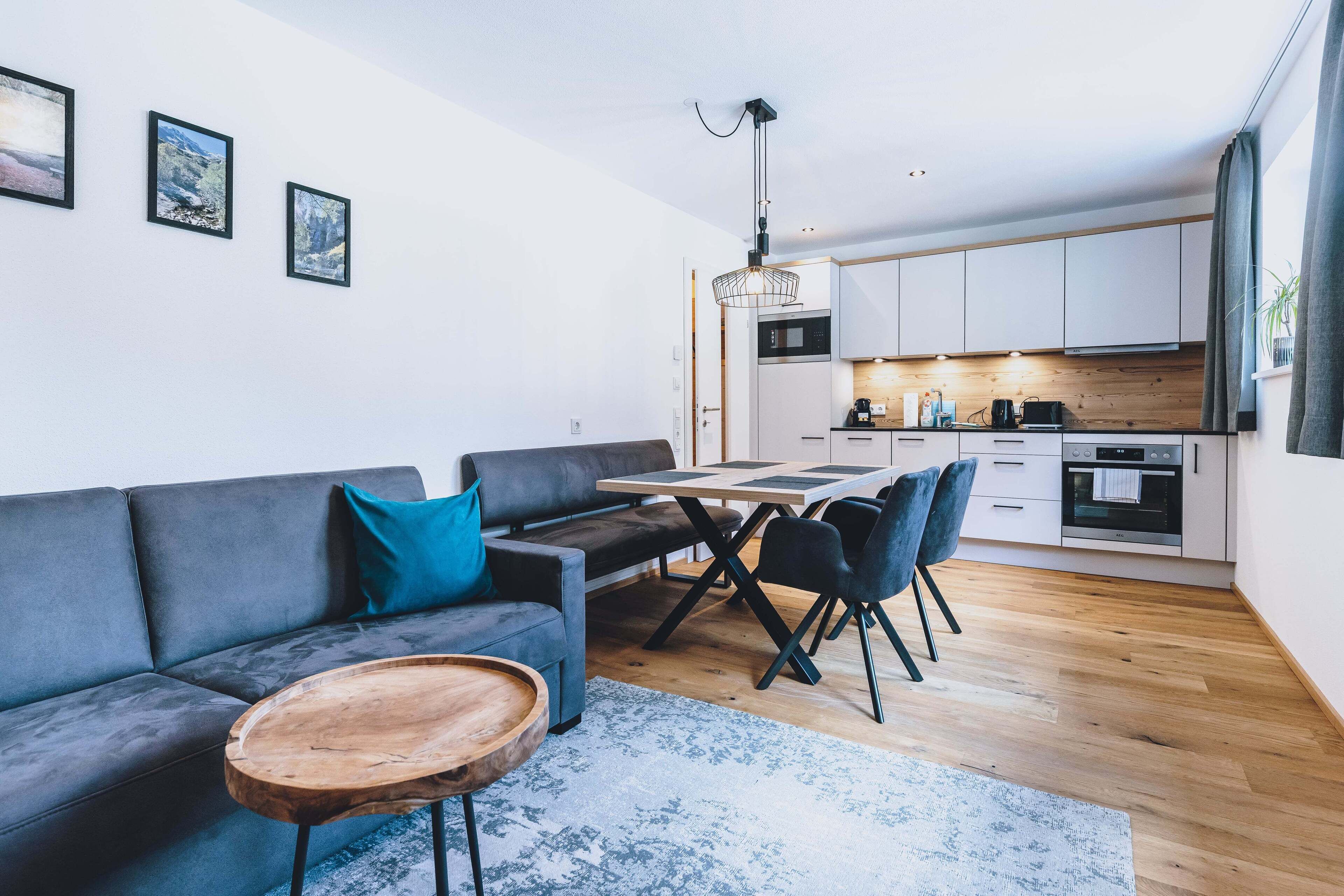 Una sala moderna con un sofá gris, una mesa de centro de madera y una zona de comedor con mesa y sillas. Se ve una cocina en el fondo con armarios blancos y una encimera de madera.