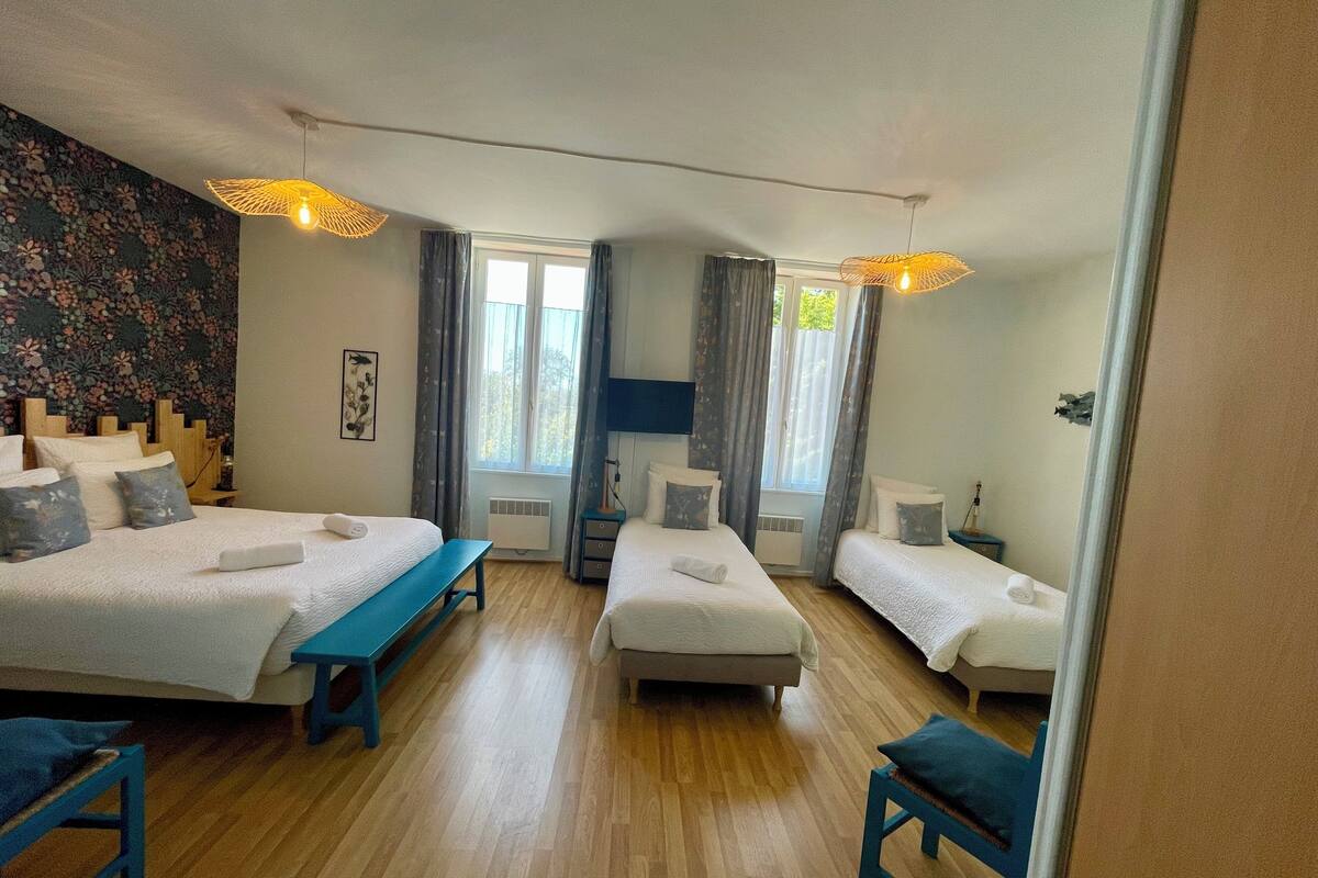 Une chambre d’hôtel avec deux lits simples, un banc, une petite table et une grande fenêtre avec des rideaux.