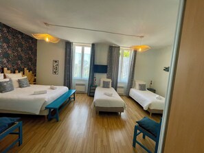 A hotel room with two single beds, a bench, a small table, and a large window with curtains.