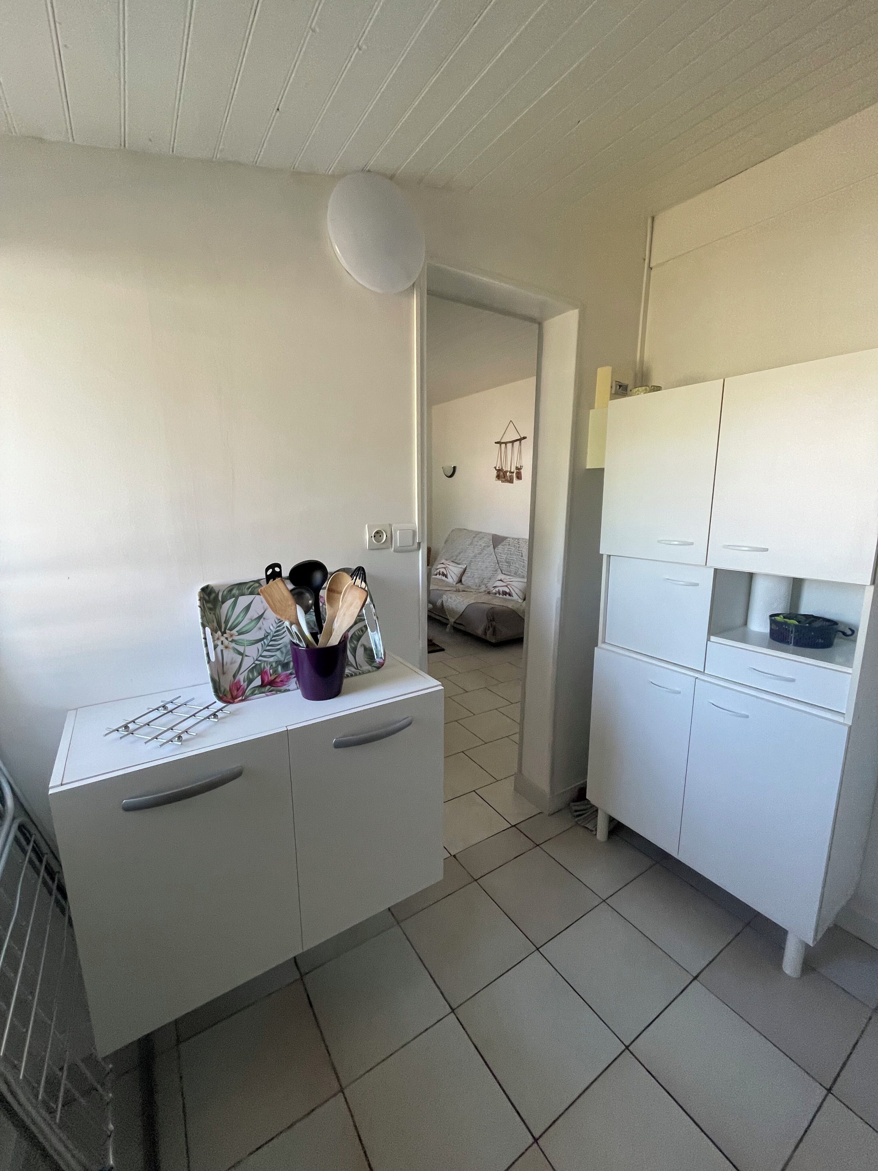 A compact kitchen with white cabinets, a countertop with kitchen utensils, and a visible bed in the adjacent room.