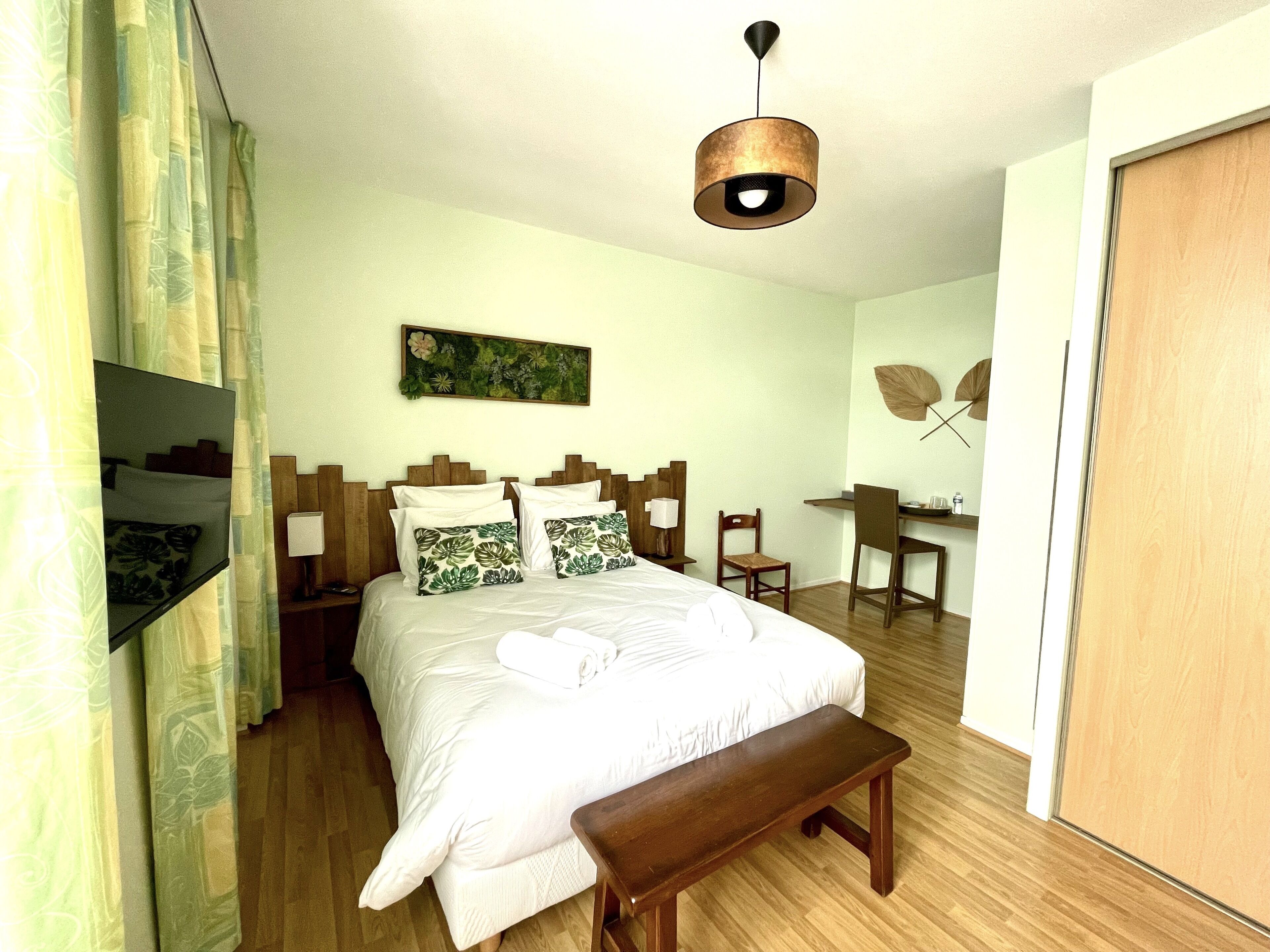 A hotel room with a bed, a desk, a chair, a bench, a TV, and a painting on the wall.