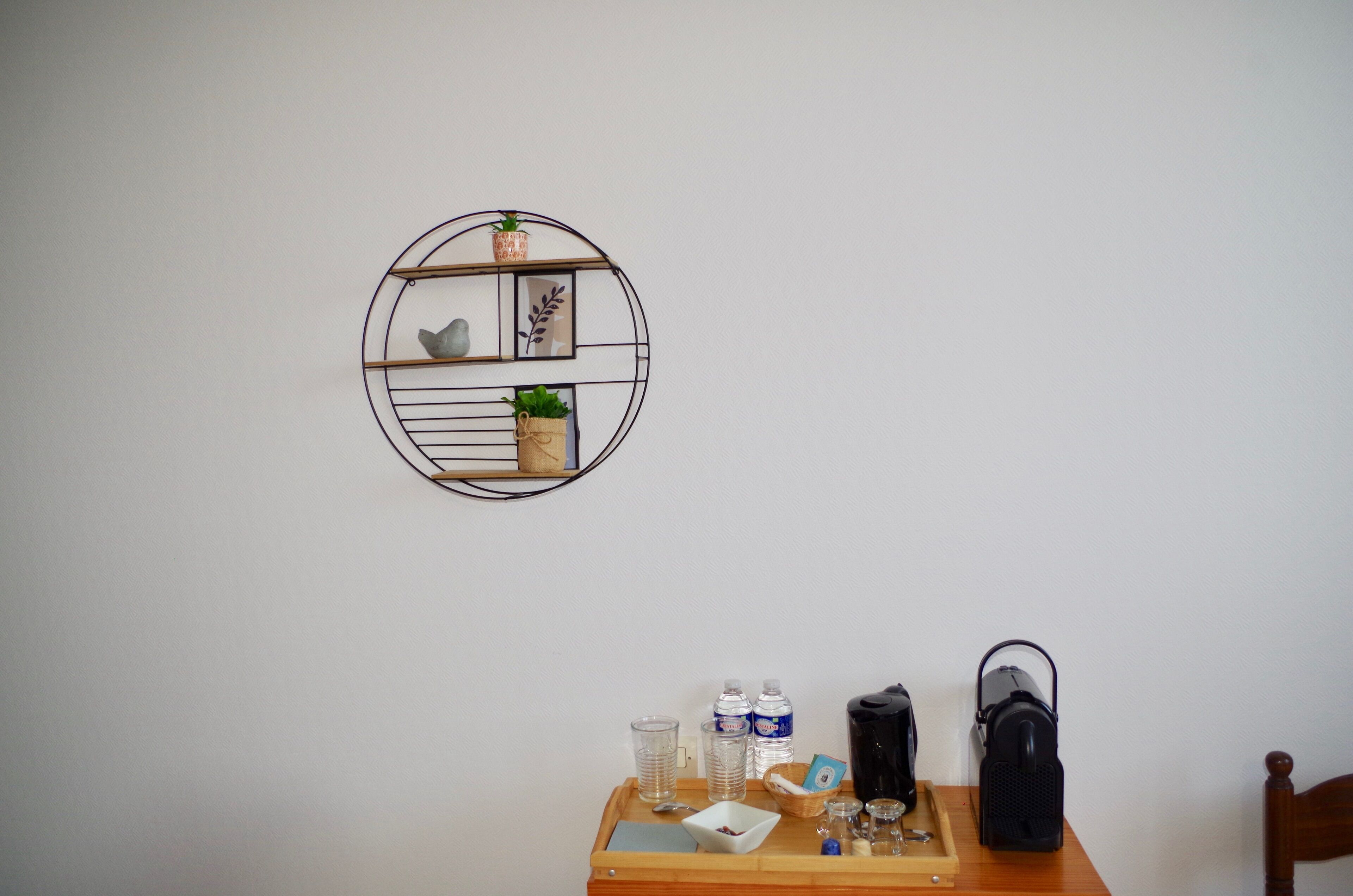 Un estante circular montado en la pared con objetos decorativos y una mesa con una bandeja que contiene una botella de agua, vasos, un pequeño cuenco y un bolso negro.