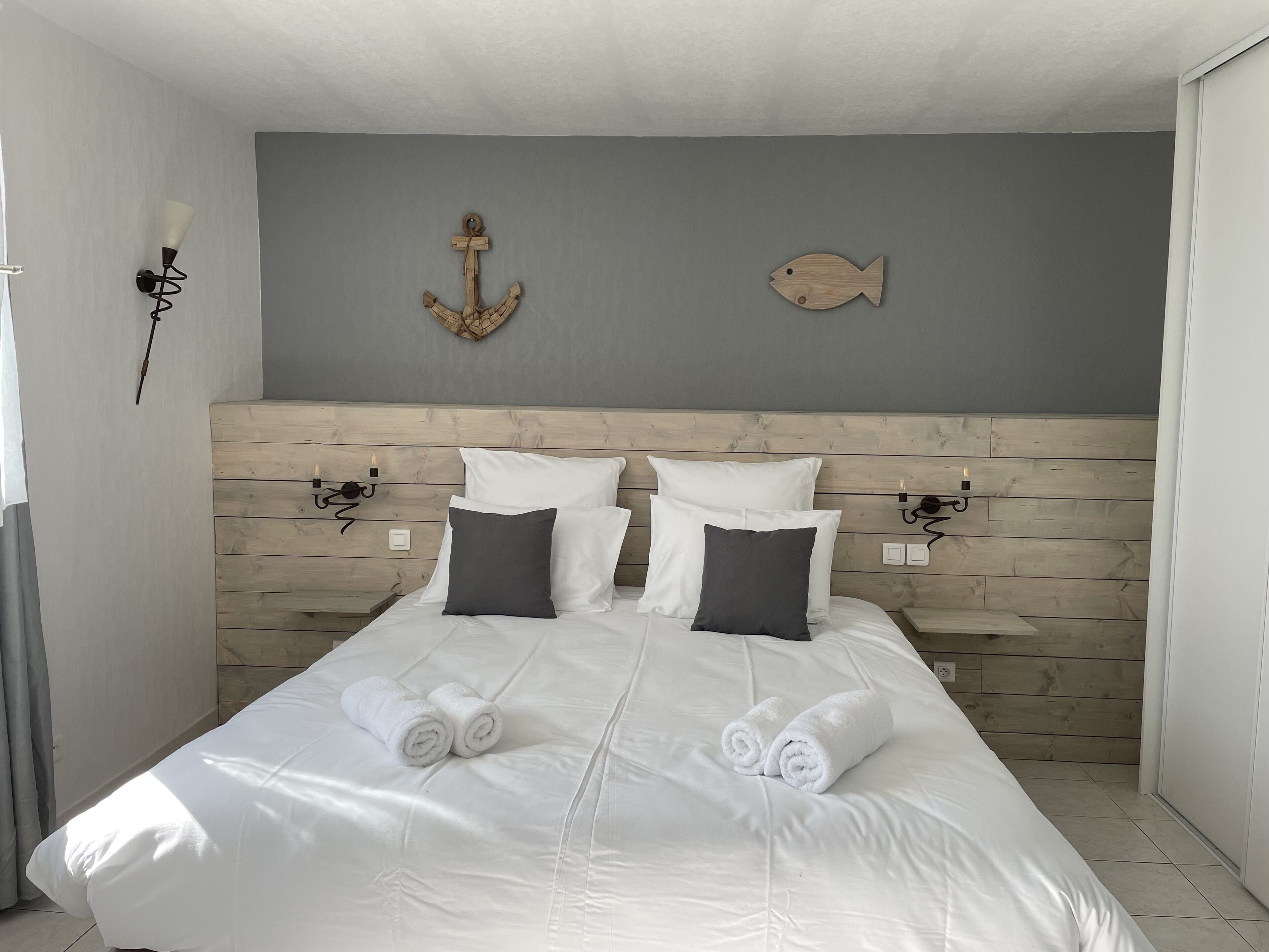 Ein ordentlich gemachtes Bett mit weißen Leinen und zwei aufgerollten Handtüchern, einem Holzkopfschild sowie Wandleuchten.