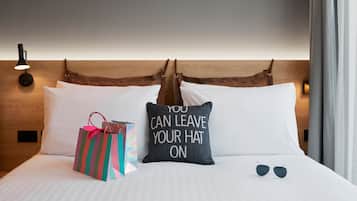 A neatly made bed with a pillow that reads "YOU CAN LEAVE YOUR HAT ON," a pair of sunglasses, and a gift bag on the bed.
