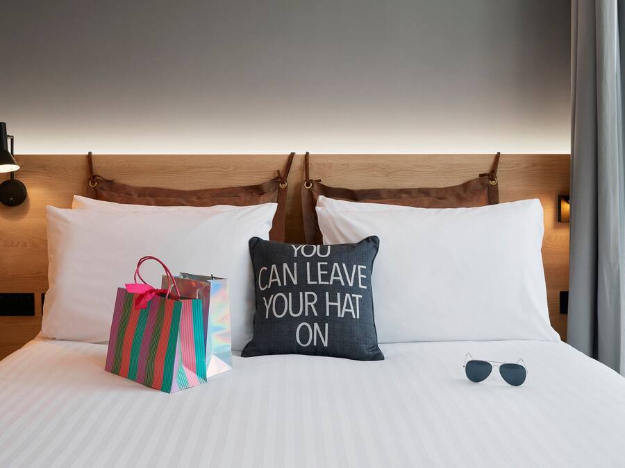 Ein ordentlich bezogenes Bett mit einem Kopfkissen, auf dem 'YOU CAN LEAVE YOUR HAT ON' steht, einer Sonnenbrille und einer Geschenktüte.