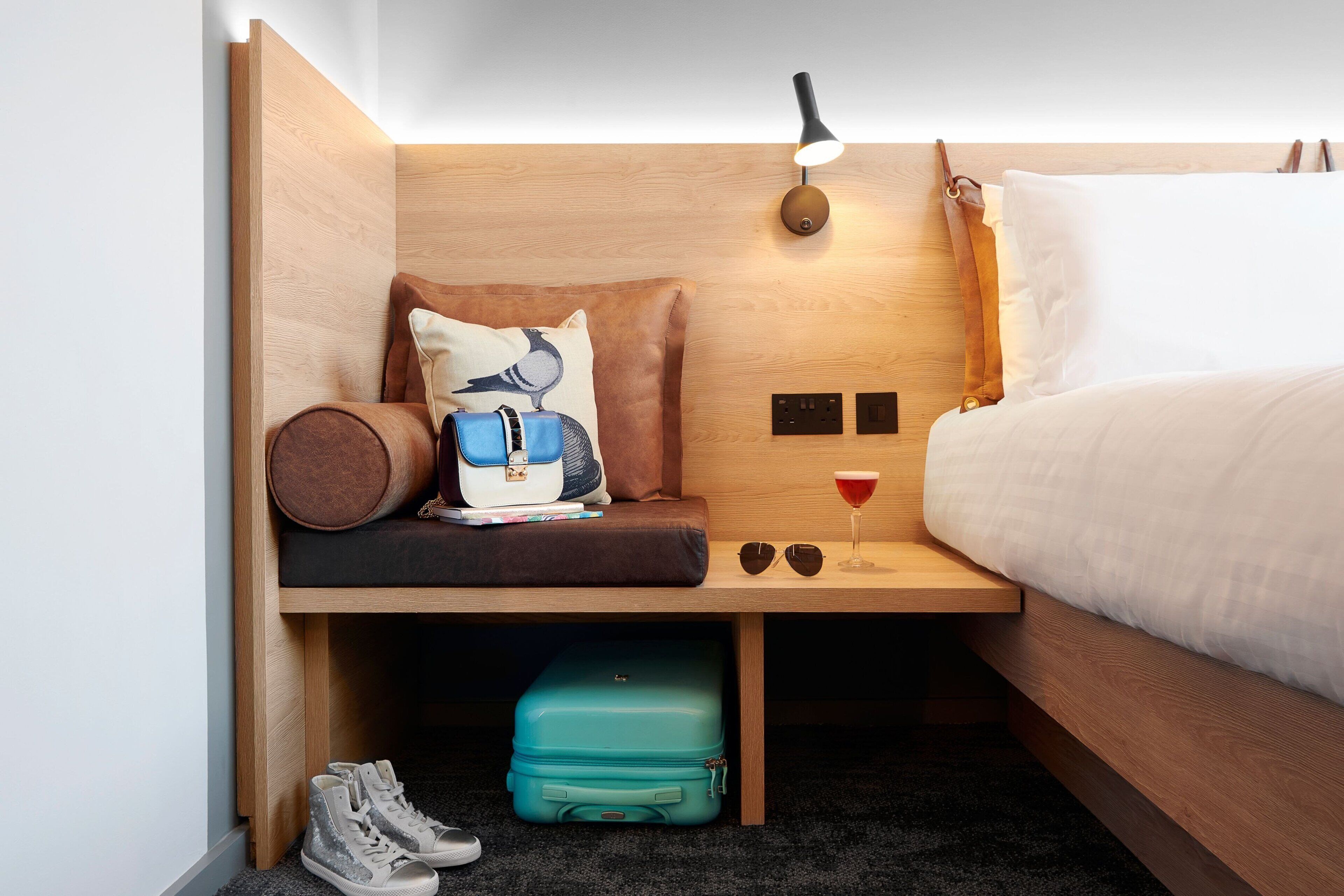 A compact room with a bed, a bench with a pillow, a small table with a glass and a drink, a suitcase, and a pair of shoes.