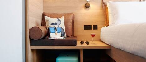 A compact room with a bed, a bench with a pillow, a small table with a glass and a drink, a suitcase, and a pair of shoes.