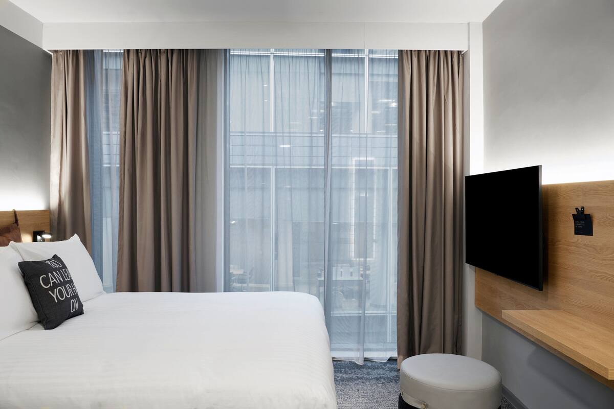 A hotel room with a large bed, a flat-screen TV mounted on the wall, a wooden headboard, and a view through sheer curtains.