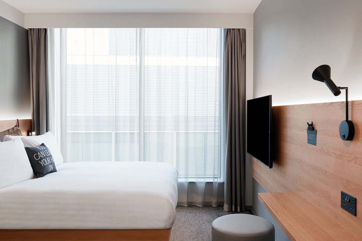 A hotel room with a bed, a TV, a desk, and a window with curtains.