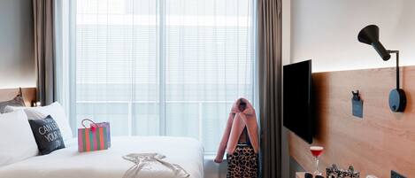 A hotel room with a bed, a TV, a desk with a lamp, a window with curtains, and a view of the city.