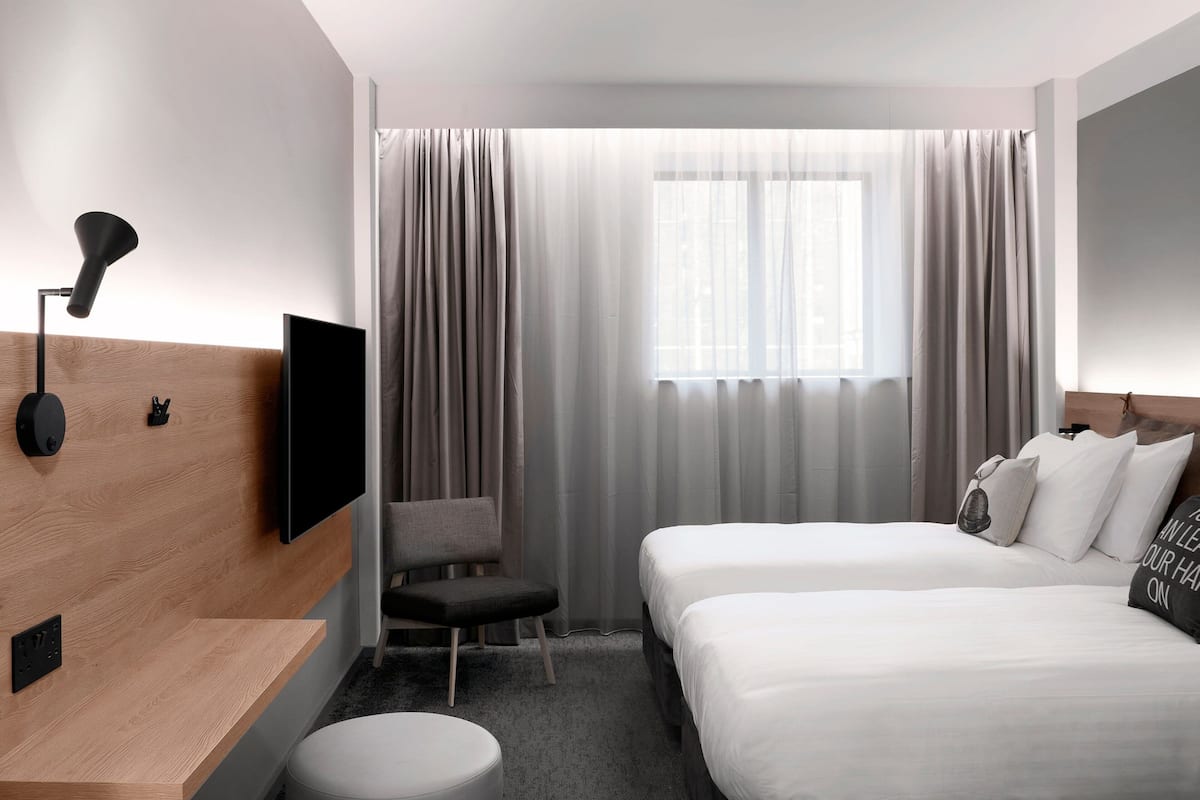 A hotel room with a wooden headboard, a flat-screen TV, a desk with a lamp, two beds, a chair, and a large window with curtains.