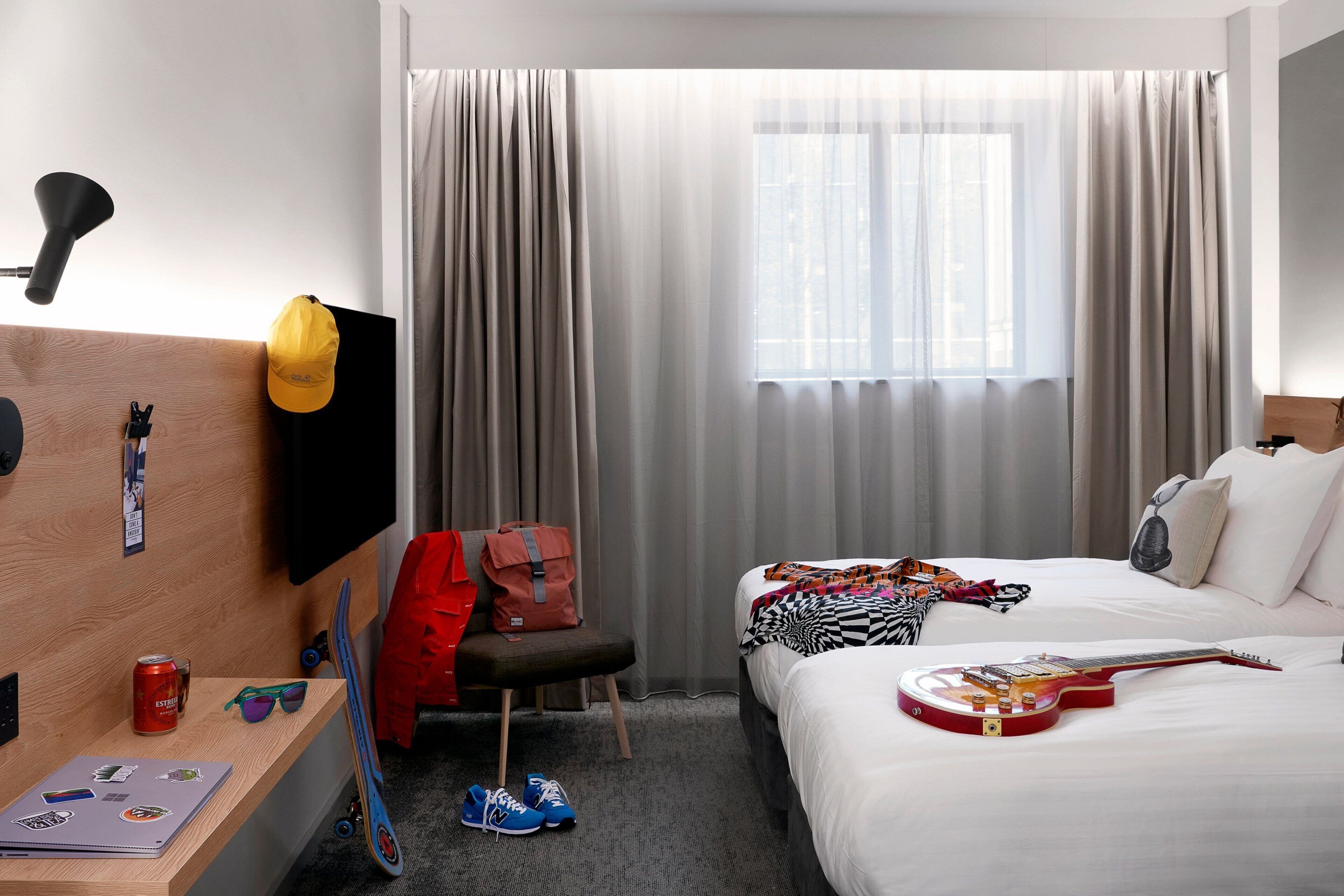 A hotel room with a bed, a desk, a chair, a TV, a window with curtains, and a guitar.