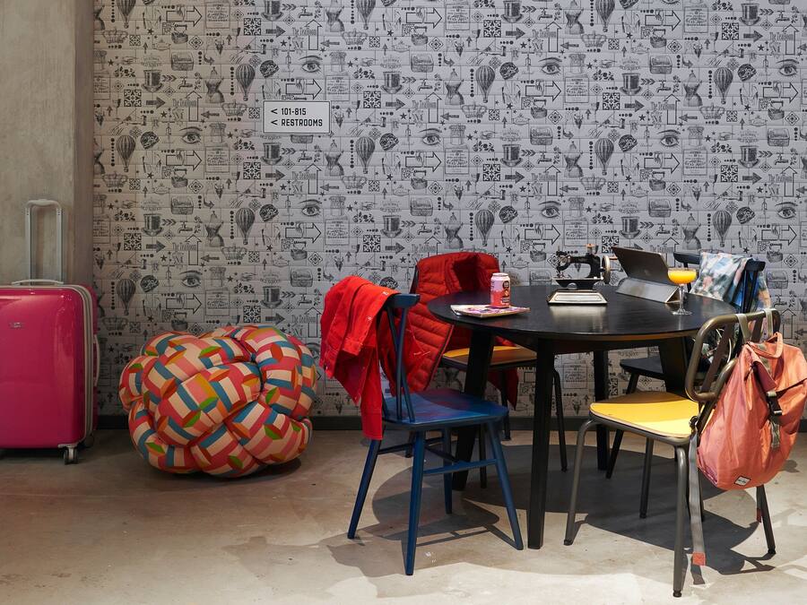 Una habitación con papel tapiz estampado, una mesa redonda, sillas, una maleta rosa, un sillón de peluche colorido y una chaqueta roja sobre una silla.