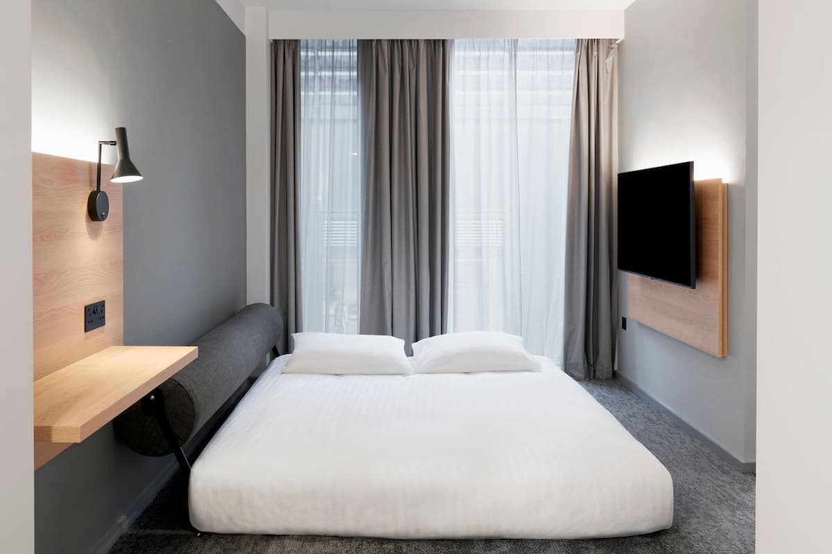A modern hotel room with a bed, a wooden shelf, a wall-mounted TV, and a window with curtains.
