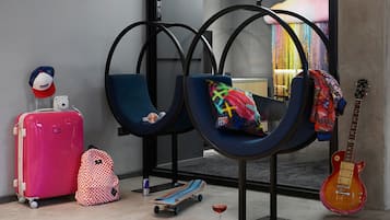A room with two blue chairs, a pink suitcase, a checkered backpack, a skateboard, and a guitar.