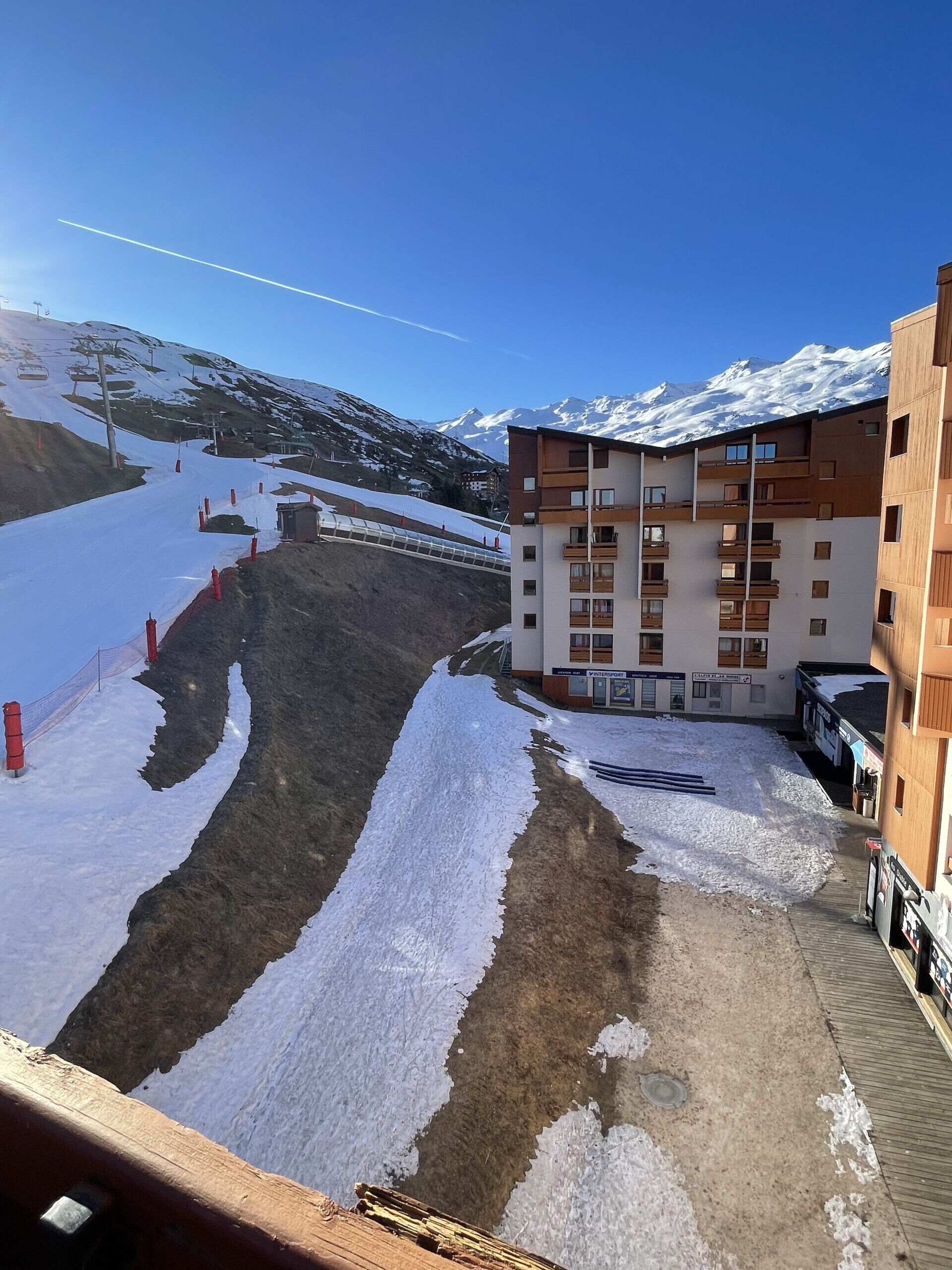 Ein modernes Gebäude mit mehreren Balkonen, im Hintergrund eine Skipiste und ein strahlend blauer Himmel.