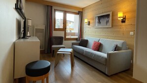 A modern living room with a sofa, a small table, and a desk. There is a window with curtains, a wall-mounted TV, and a wooden panel wall.