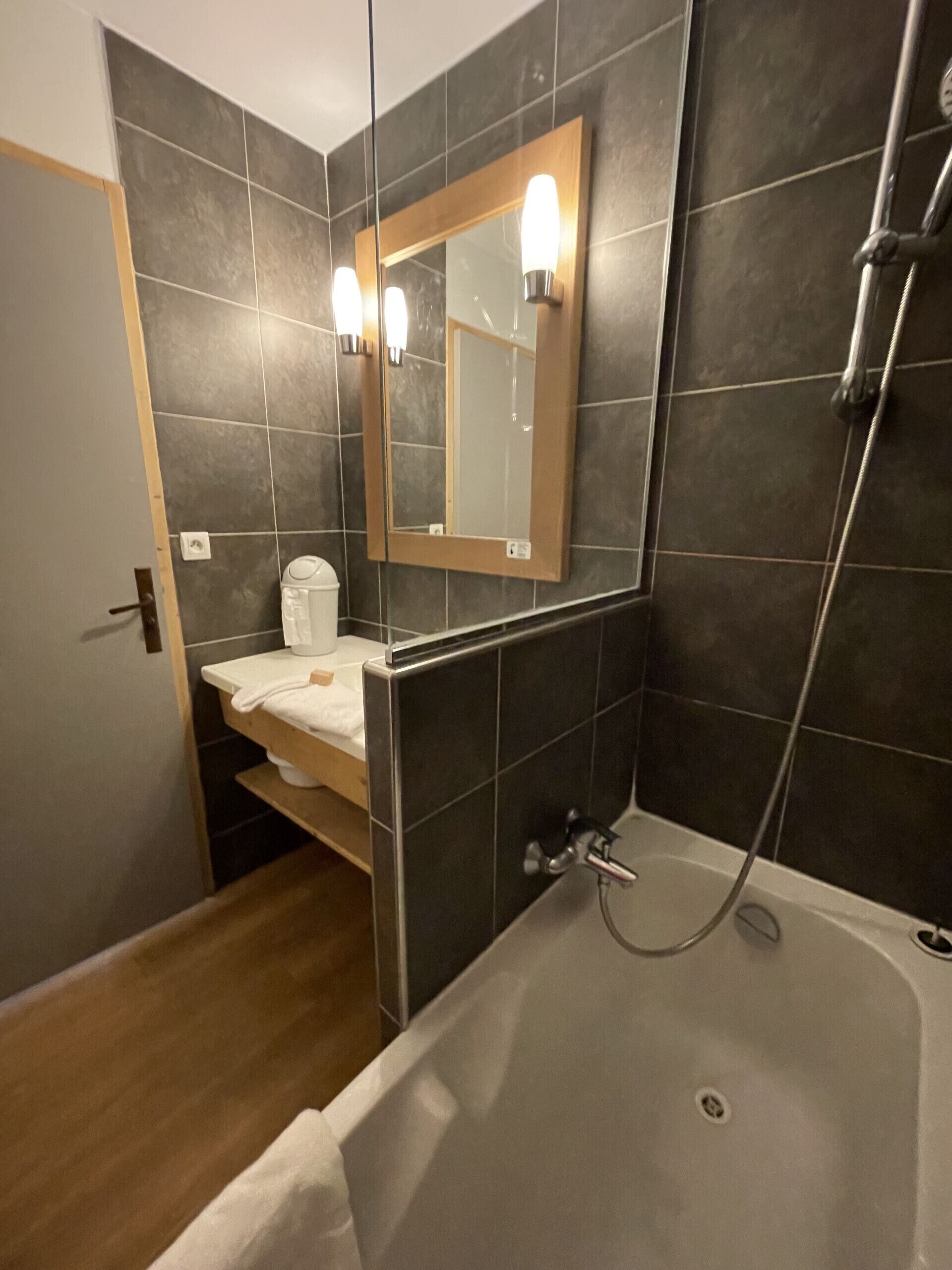 A modern bathroom with dark tiles, a wooden door, a sink with a towel, a mirror with lights, and a shower area.