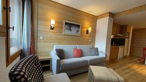 A living room with a sofa, a chair, and a small table. There is a wooden wall with a picture frame and wall-mounted lights. A kitchen is visible in the background.