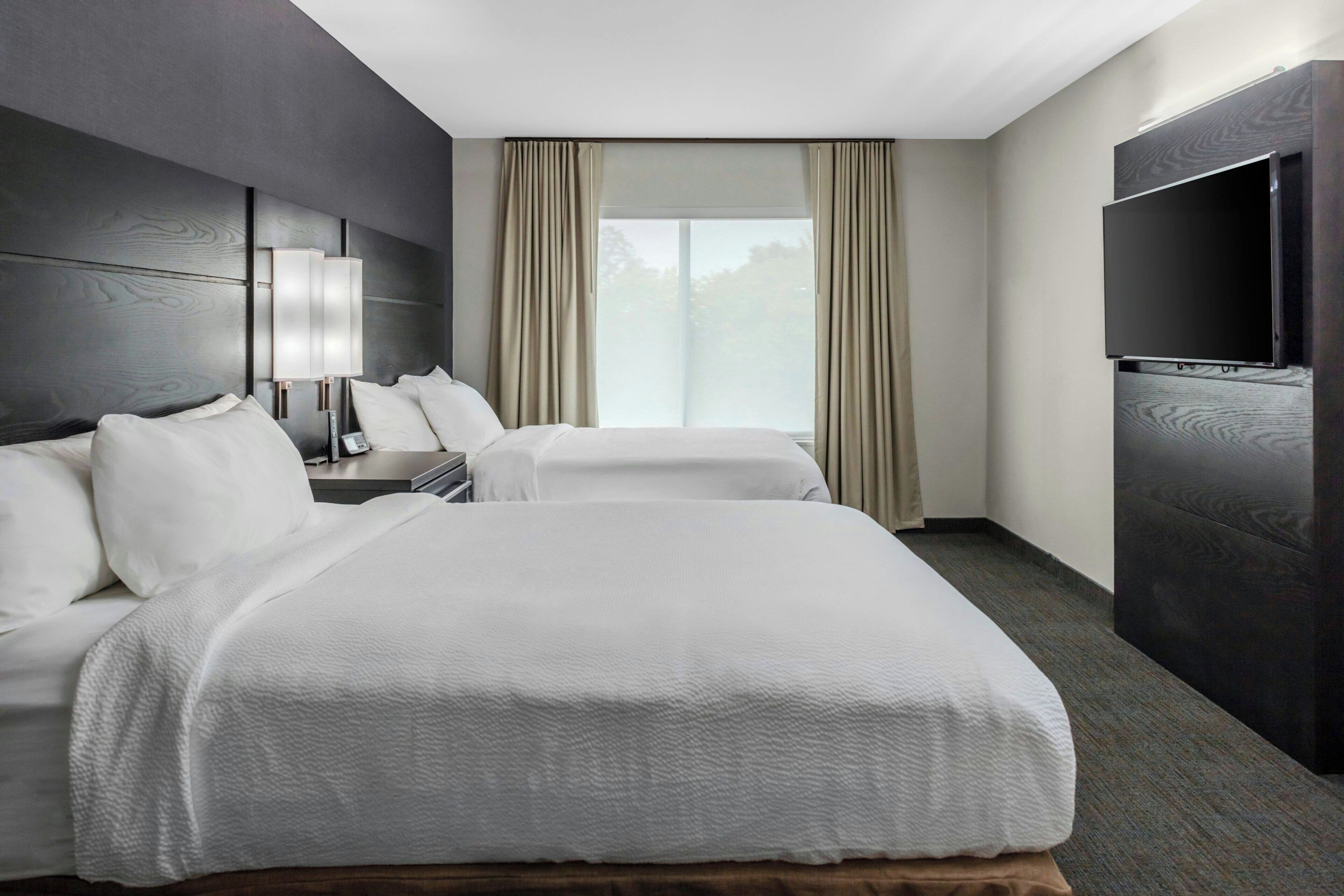 Habitación de hotel con dos camas, una televisión de pantalla plana, una mesita de noche y una ventana con cortinas.