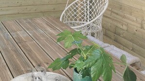 Une terrasse en bois agrémentée de guirlandes lumineuses, d’un fauteuil suspendu et d’une table sur laquelle sont posés un pot de fleurs et des bocaux en verre contenant des pailles.