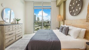 Une chambre moderne avec un grand lit, une commode, un miroir et une vue sur la ville par une grande fenêtre.