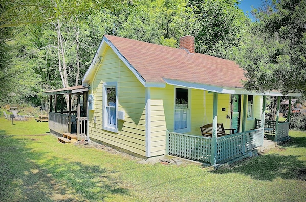 A small, single-story house with a porch, surrounded by a grassy yard and trees.