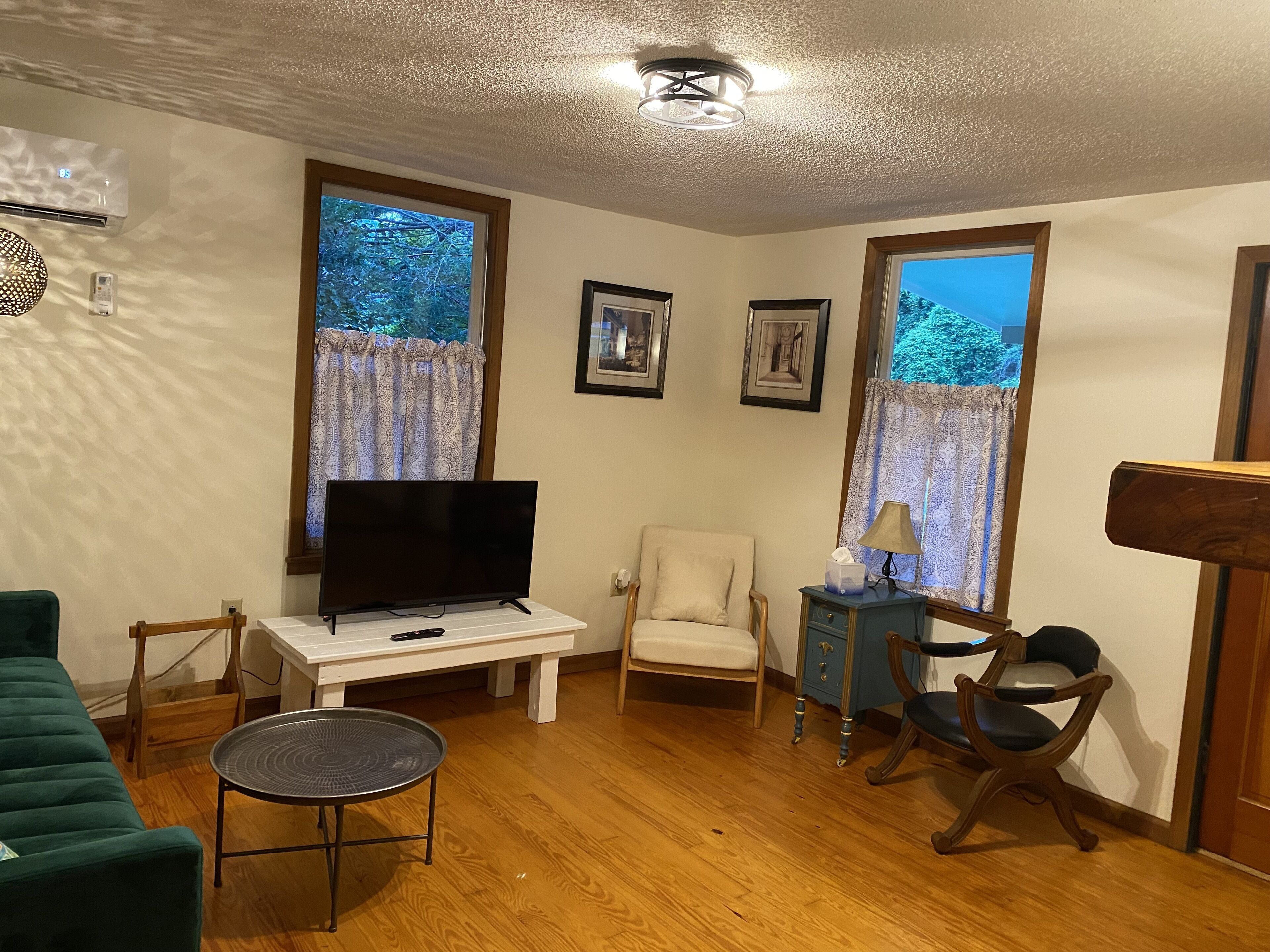 A living room with a flat-screen TV on a white table, a green sofa, a small round table, and a wooden floor.