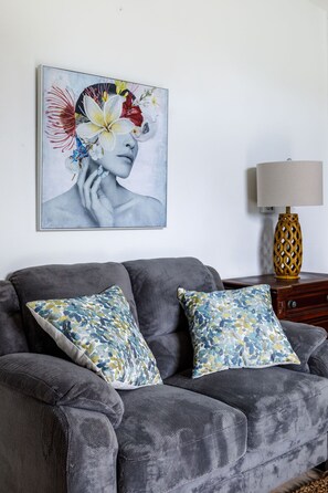 A grey sofa with floral-patterned cushions, a wall-mounted painting, and a table lamp with a gold base.