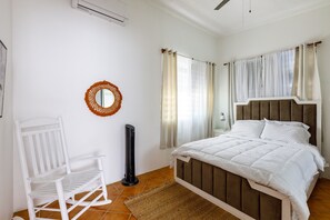 A bedroom with a bed, a chair, a standing heater, and a mirror on the wall.