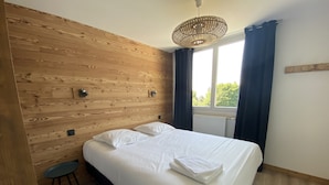 A bedroom with a wooden headboard, a bed with white linens, a small stool, and a window with curtains. - 6/8 pers apartment in the heart of the Chartreuse at the foot of the ski slopes (Sarcenas)