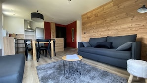 A modern living room with a dark grey sofa, a wooden dining table with chairs, and a kitchen area in the background. - 6/8 pers apartment in the heart of the Chartreuse at the foot of the ski slopes (Sarcenas)