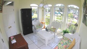 Une pièce baignée de lumière, avec des meubles en osier peints en blanc, une table en bois et un buffet noir.
