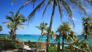 Eine Holzterrasse mit zwei gestreiften Stühlen, die auf ein türkisblaues Meer ausgerichtet sind, umgeben von Palmen.