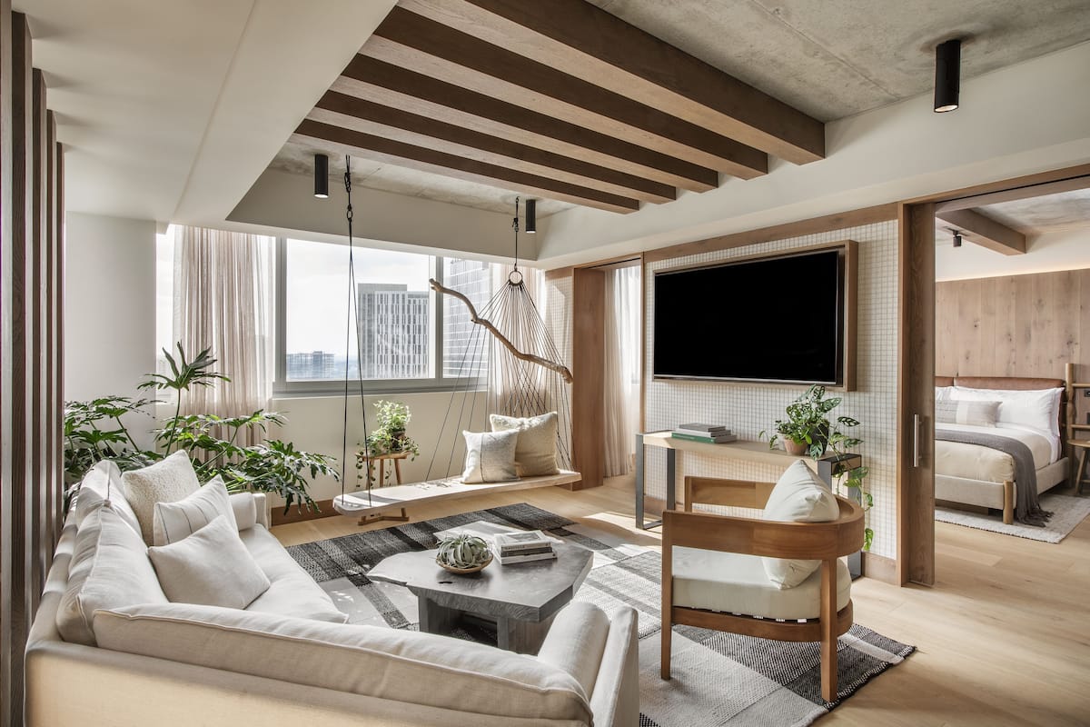 Ein modernes Wohnzimmer mit Sofa, Couchtisch und Hängekorbsessel. An der Wand ist ein Fernseher befestigt, im Hintergrund steht ein Bett.