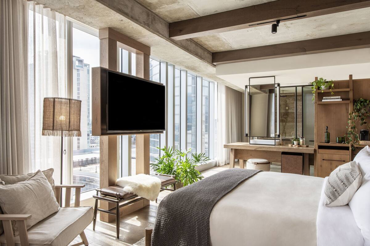 Ein modernes Hotelzimmer mit einem großen Bett, einem Schreibtisch, einem Fernseher und Blick auf die Stadt durch große Fenster.