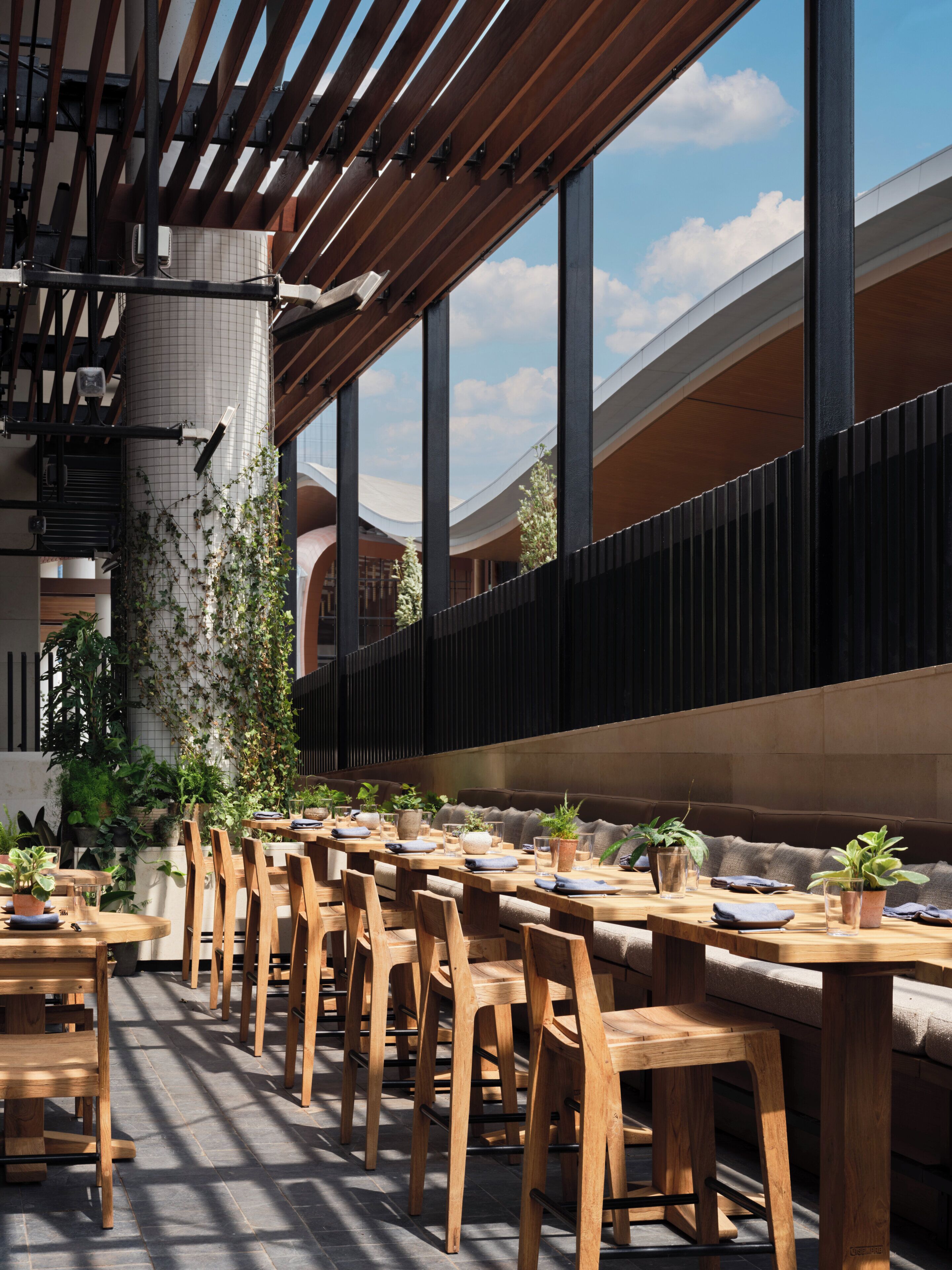 A modern restaurant with wooden tables and chairs, potted plants, and a view of the sky through large windows.
