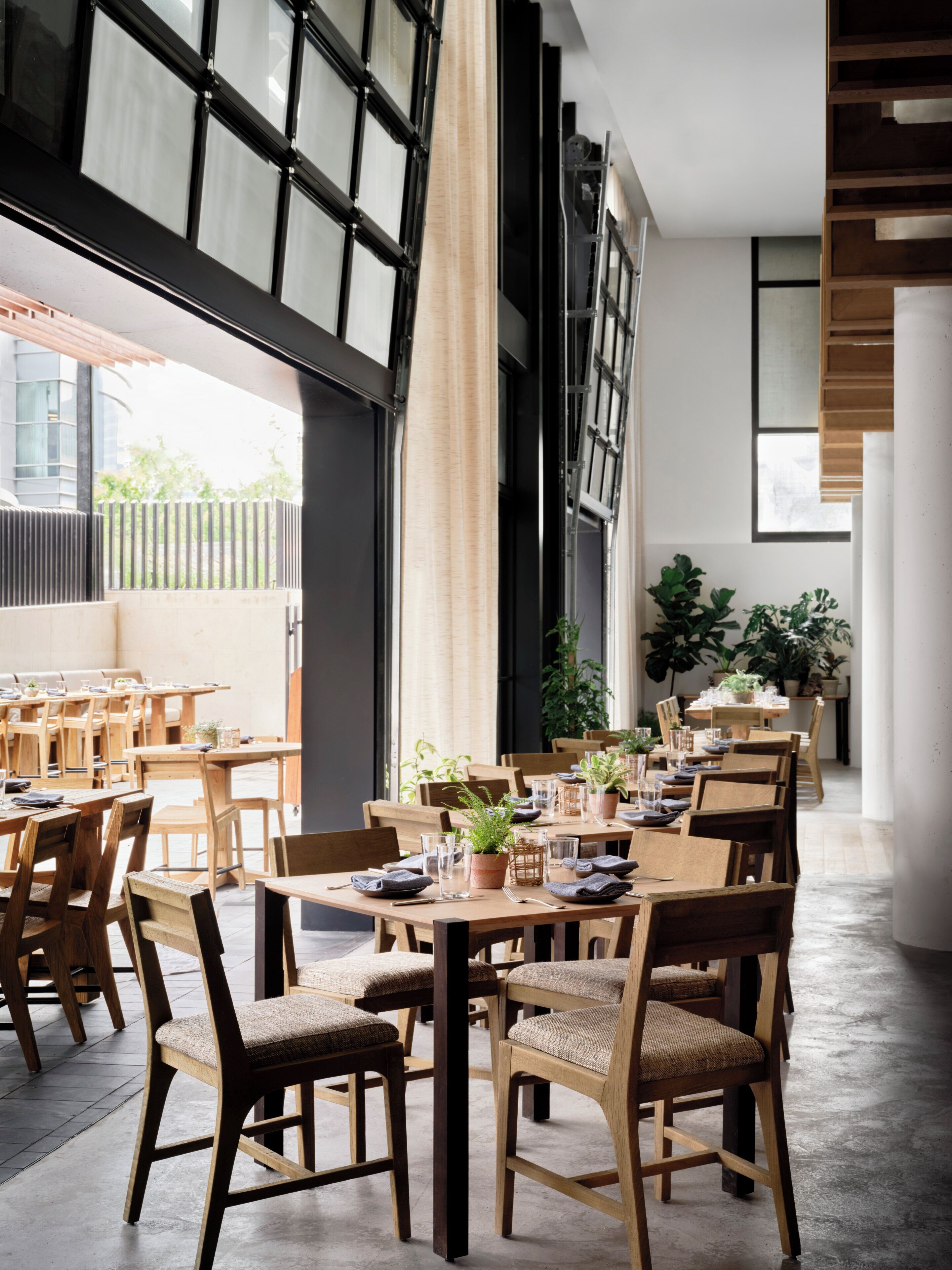 A restaurant with wooden tables and chairs, large windows, and indoor plants.