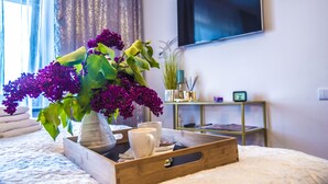 Puisesta alustasta on aseteltu hotellihuoneen yöpöydälle maljakko, jossa on violetteja kukkia, kaksi valkoista kuppia ja pieni kello.