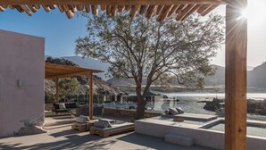 Ein sonnendurchfluteter Außenbereich mit Pergola, einem Baum und Blick auf ein Gewässer und Berge.
