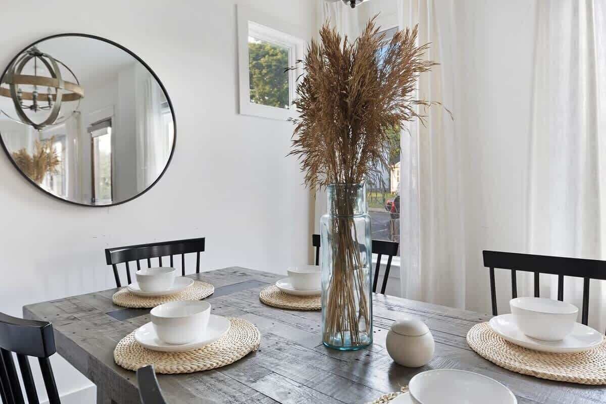 Une table à manger dressée avec des assiettes et des bols blancs, un vase contenant des plantes séchées et un miroir rond accroché au mur.