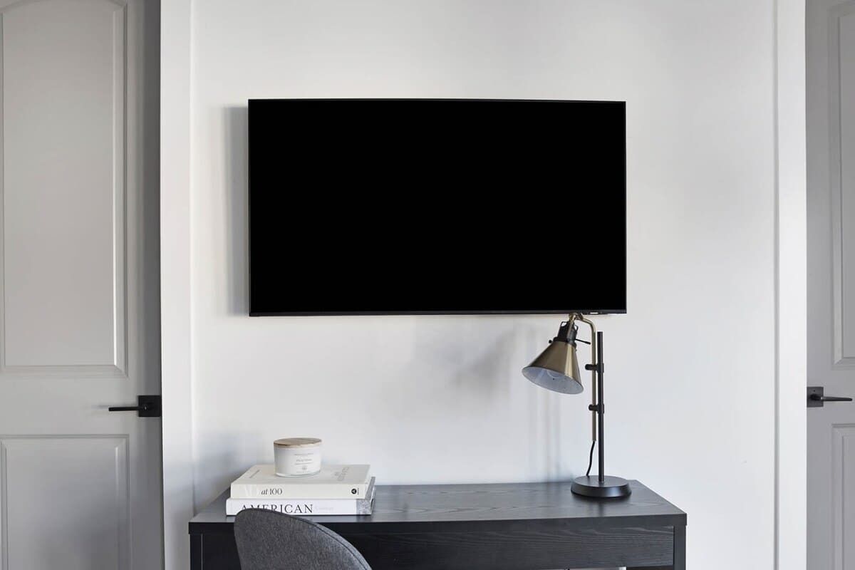 A room with a flat-screen TV mounted on the wall, a desk with a lamp, and a chair.