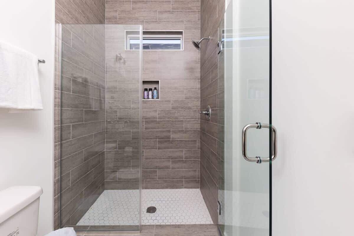 Ducha con paredes de vidrio, azulejos grises, un pequeño estante con artículos de aseo y una toalla blanca en la pared.