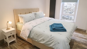 Una cama bien hecha con una colcha azul estampada con flores, una pila de toallas azules dobladas, una lámpara de noche y una ventana con vistas a terreno rocoso.