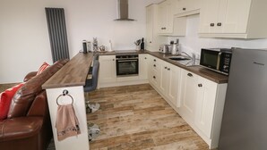 Cocina compacta con cubierta de madera, armarios blancos, horno integrado, microondas y zona de estar con sofá de cuero marrón.