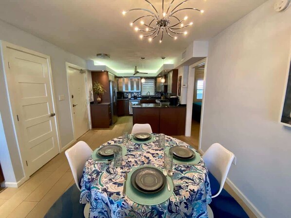 Comedor con una mesa redonda dispuesta para cuatro, una cocina al fondo y un ventilador de techo.