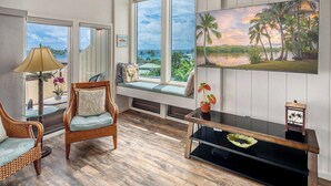 Une pièce dotée d’une grande fenêtre donnant sur une plage, d’un fauteuil en osier, d’une table avec une lampe et d’un tableau représentant une scène de plage.