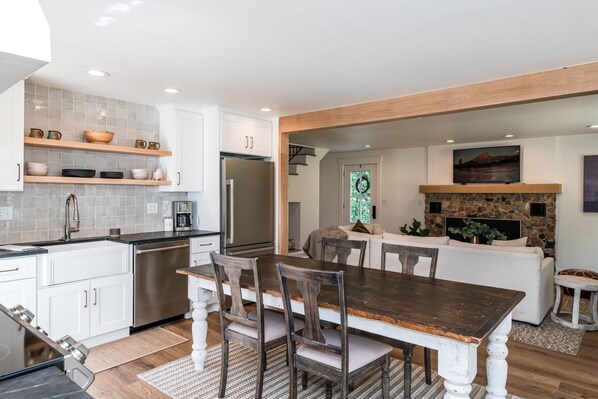 A modern kitchen with white cabinetry, a wooden dining table, and a stone fireplace.